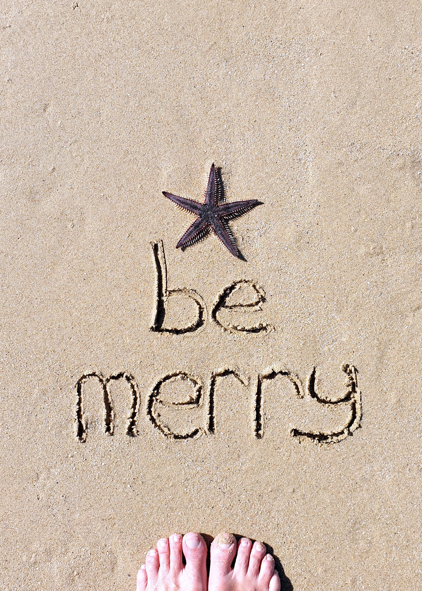 Be merry,Christmas text words written on sand with starfish and feet in frame